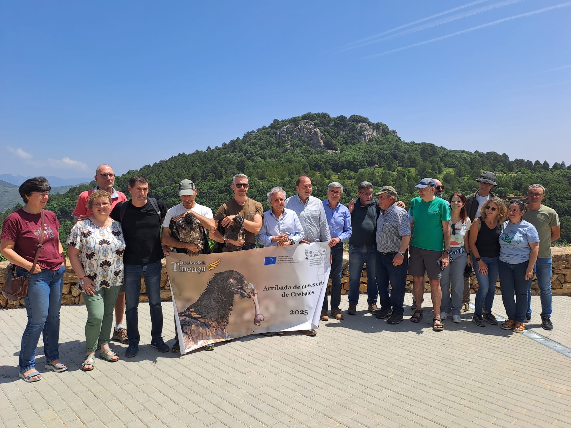A froup of people holding a banner with a Bearded vulture printed on. Two people hold one young bearded vulture each. Mountains in the background